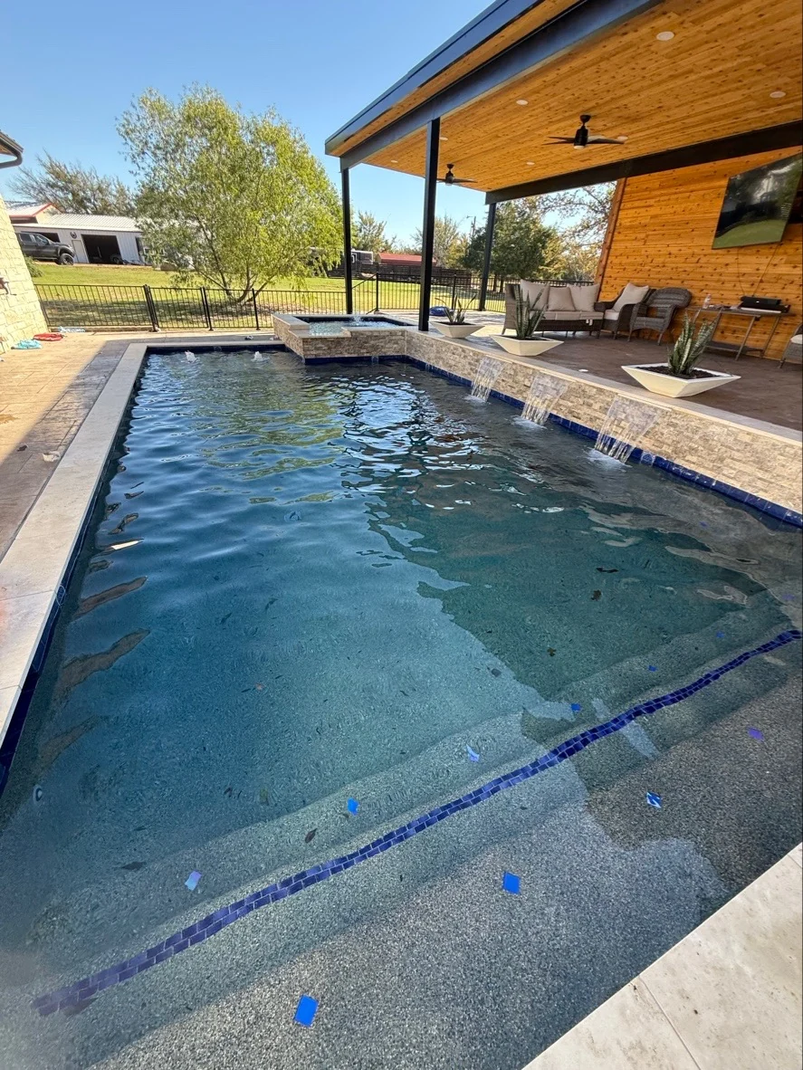 Covered poolside outdoor living space with wood ceiling, waterfall detail, and seating by Averra Pools and Outdoor Living